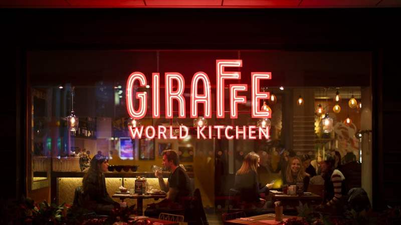 cửa sổ kính từ bên ngoài nhìn vào nhà hàng với biển hiệu Giraffe World Kitchen lớn trong đèn neon đỏ