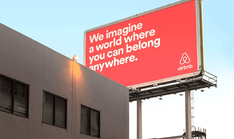 một bảng quảng cáo airbnb màu hồng san hô và dòng chữ: chúng tôi tưởng tượng ra một thế giới mà bạn có thể thuộc về ở bất cứ đâu.