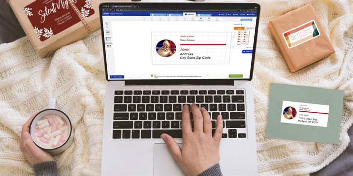 9 mẹo hay nhất cho việc gửi và gửi thư trong ngày lễ 2 Góc nhìn người đầu tiên về một vòng được phủ trong một chiếc chăn ấm áp với một chiếc máy tính xách tay trên chân của họ. Người đó cầm ca cao với kẹo dẻo bằng một tay và tay kia đang thiết kế nhãn địa chỉ Best Print trên máy tính xách tay của họ. Hai gói và một phong bì đựng thiệp lễ được trải rộng với các nhãn Best Print được làm bằng thiết kế mẫu Best Print miễn phí.