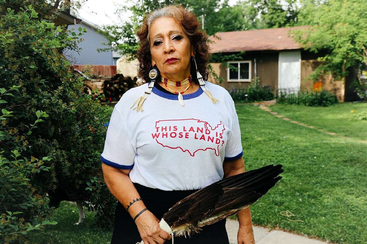 Một phụ nữ bản địa đứng trong sân mặc áo sơ mi đọc, "Đất này là đất của ai?"
