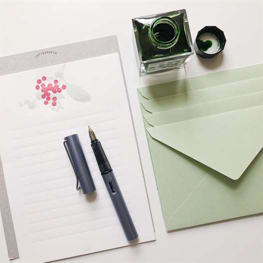 Một cây bút máy nằm trên sổ ghi chú.  Một lọ mực màu xanh lá cây ở trên cùng của hình ảnh với các phong bì màu xanh lá cây ở bên phải.