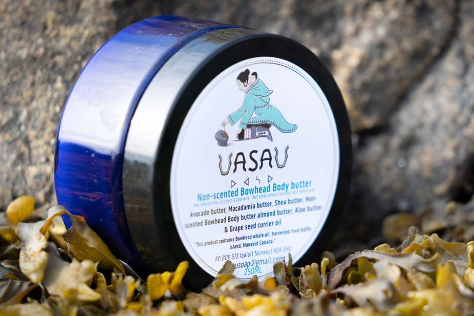 Cận cảnh một lọ đựng bơ xà phòng UasaU hình tròn đặt trên một tảng đá có địa y xung quanh.  Nhãn có biểu tượng xà phòng UasaU là hình vẽ một người mẹ Inuit đang ôm đứa con của mình. 