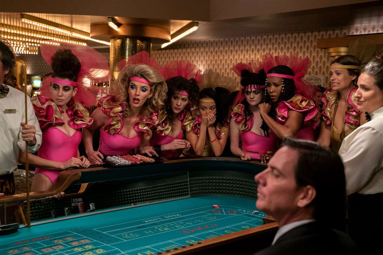 Một bức ảnh từ GLOW của Netflix với các đô vật tụ tập quanh bàn craps ở Vegas trong khi mặc trang phục sân khấu màu hồng và vàng xếp nếp.  Cherry Bang vừa tung xúc xắc, và tất cả đều đang chờ đợi xem cô ấy có thắng hay không. 