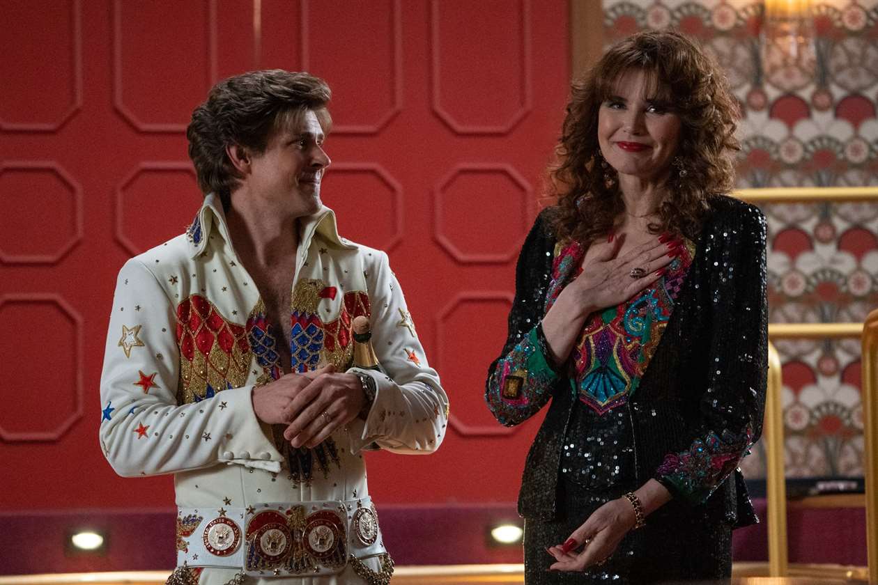 Sandy Devereaux St. Clair (Geena Davis) và Bash trong Phần 3 của GLOW.  Bash, mặc một bộ đồ có họa tiết giống với bộ đồ mà Evel Knievel sẽ mặc, đang nhìn Geena, người cũng đang mặc một bộ váy đính sequin.  Sandy đặt tay lên trái tim và mỉm cười như thể ai đó vừa nói điều gì đó tốt đẹp.