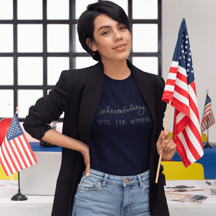 Người phụ nữ đứng trước bàn làm việc với những lá cờ Mỹ.  Áo phông của cô ấy đọc "bầu cử cho phụ nữ"