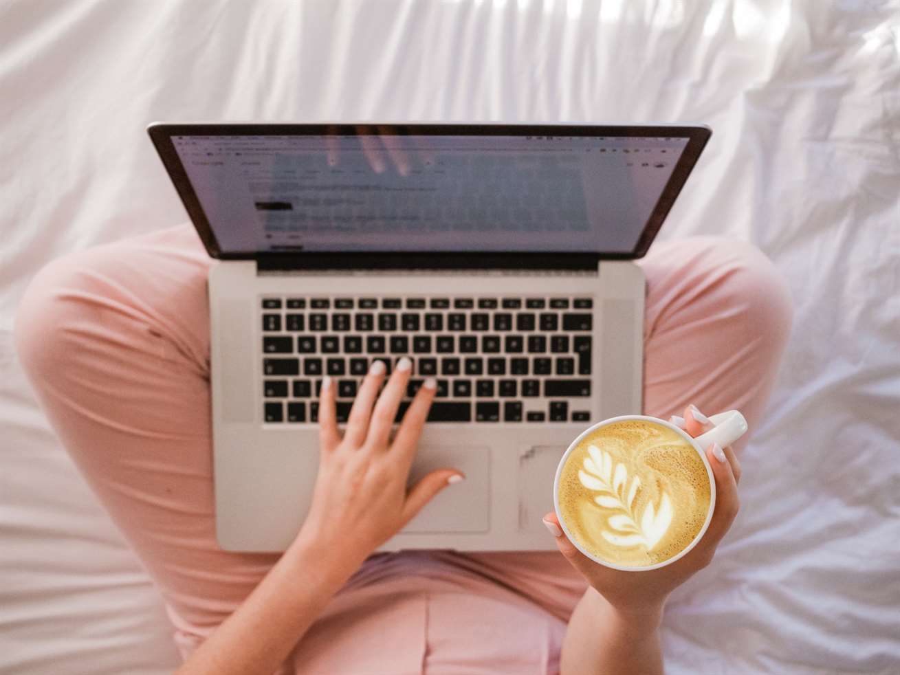 Một người ngồi xếp bằng trên giường, đặt máy tính xách tay lên đùi và cầm một ly cà phê pha cà phê